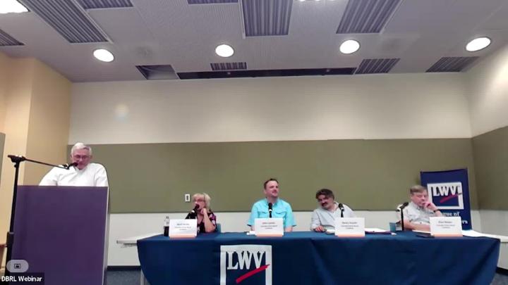 Four school board candidates sit at a table with the moderator at a podium to the left.
