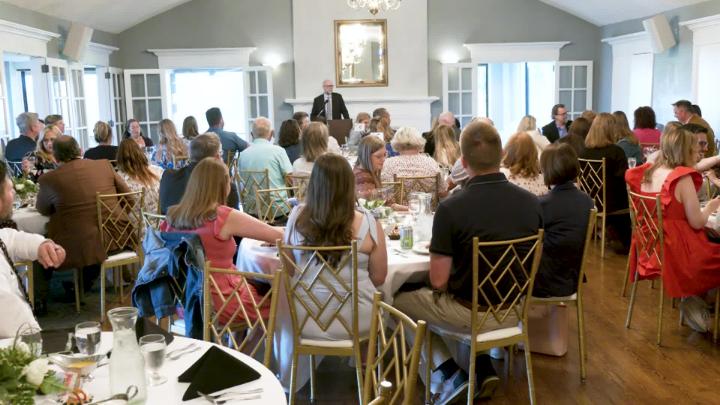 A banquet room filled with people listening to a person presenting from a podium in the front.